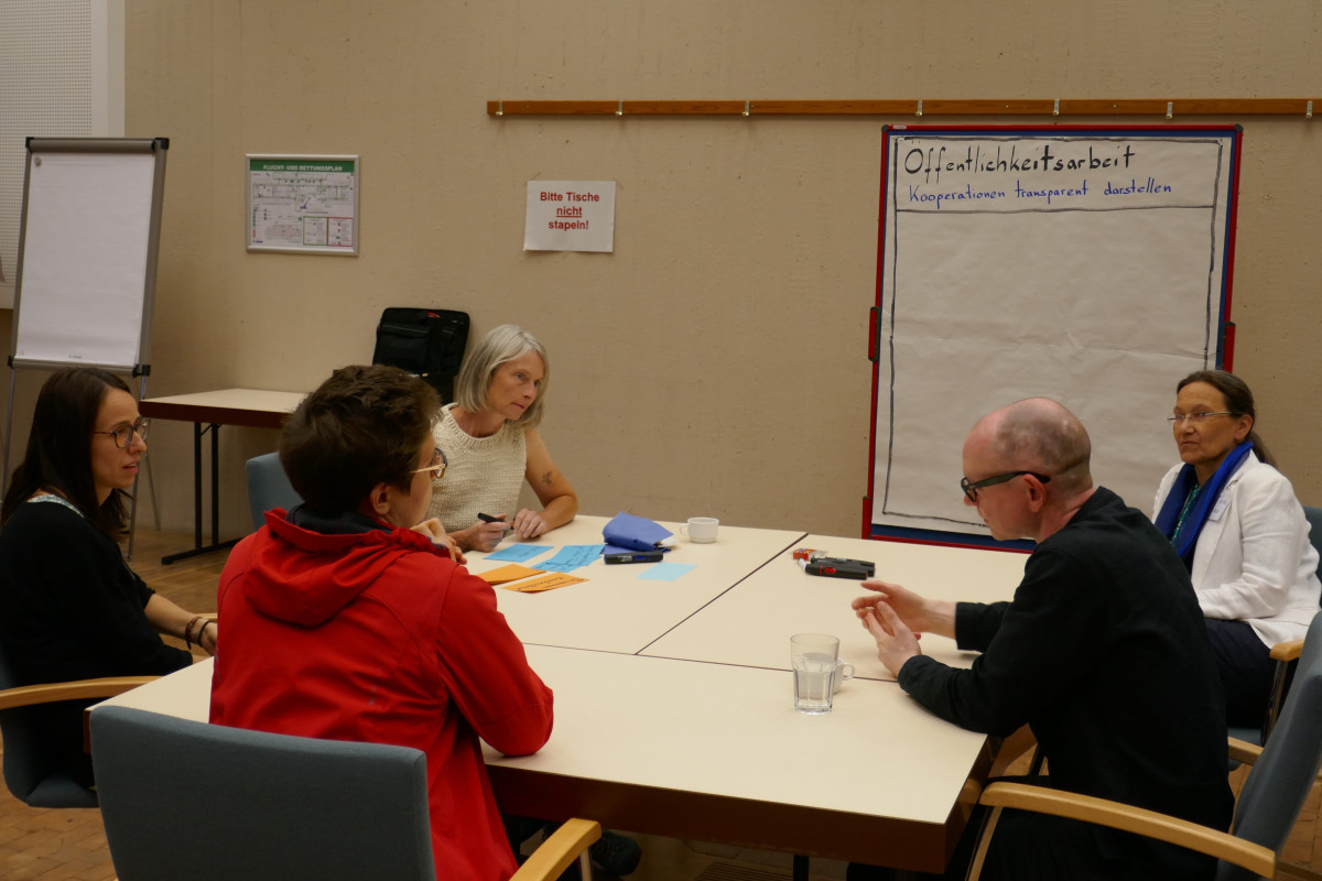 Projektmanagerinnen sitzen gemeinsam um einen Tisch herum, im Hintergrund ein Whiteboard auf dem Öffentlichkeitsarbeit draufsteht