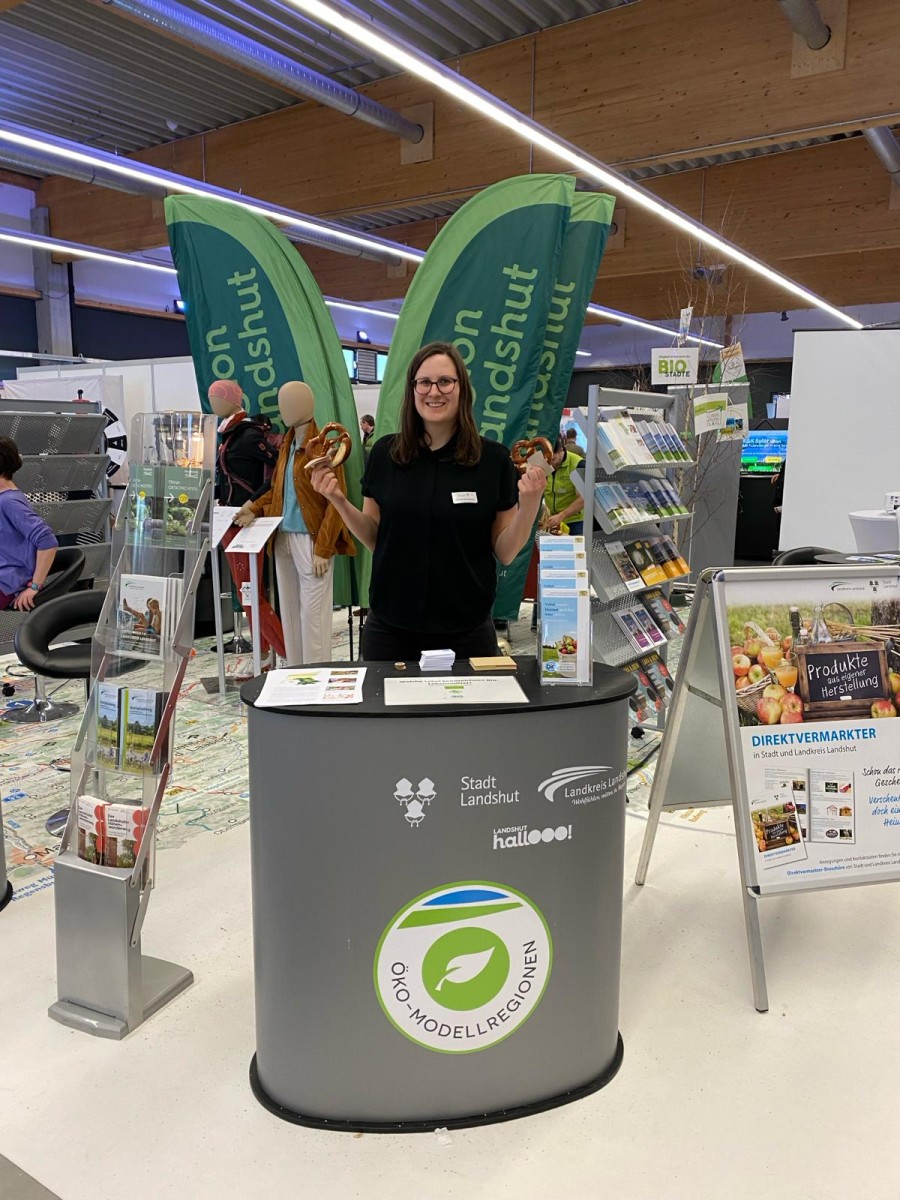 ÖMR-Managerin Veronika Stanglmayr steht hinter ihrem Stand auf der Umweltmesse in Landshut und hält zwei Brezen in die Luft.