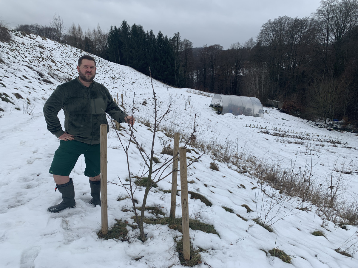 Gemüsegärtner Hans-Joachim steht im Schnee an seinem Feld und zeigt die Beerensträucher, die er im Sommer ernten wird.